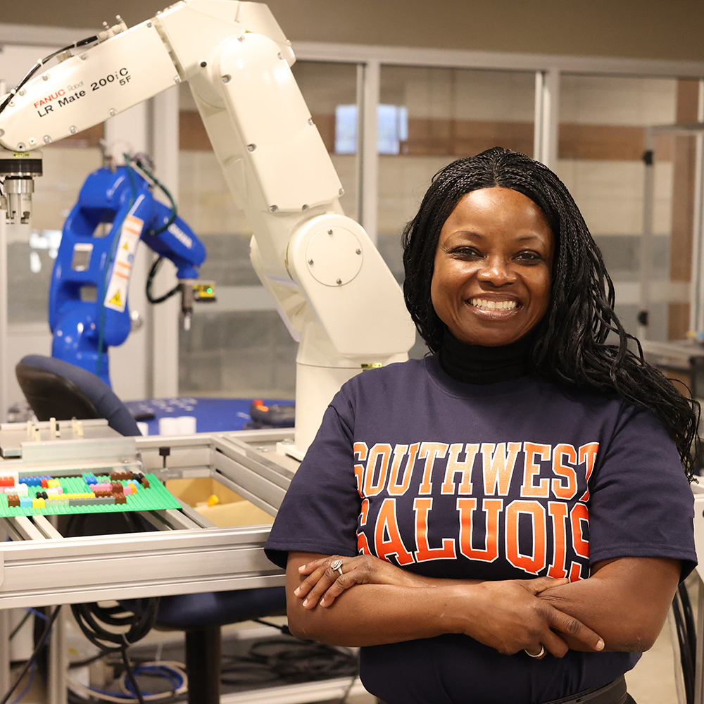 Smiling woman in a "Southwest Saluquis" shirt stands confidently in a robotics lab, with robotic arms in the background.