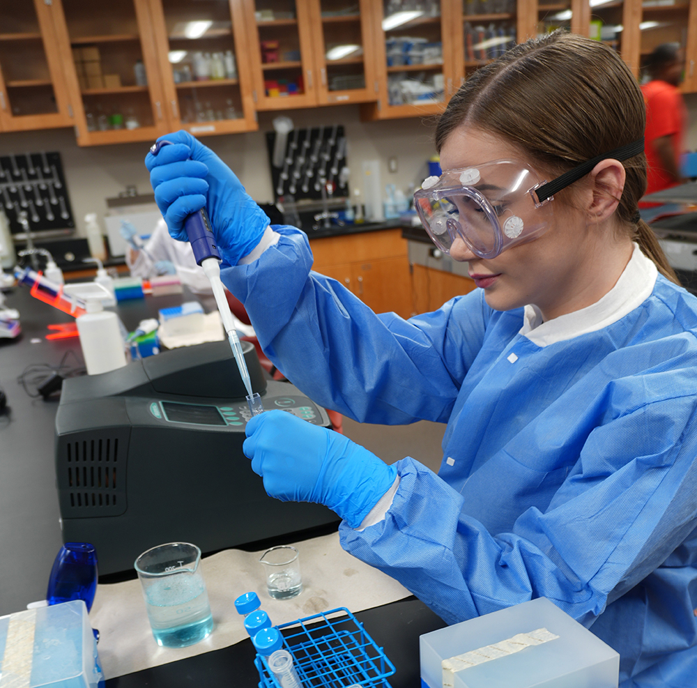 A scientist in blue protective gear uses a pipette in a lab. She wears goggles and gloves, focused and precise. Lab equipment and shelves are visible.