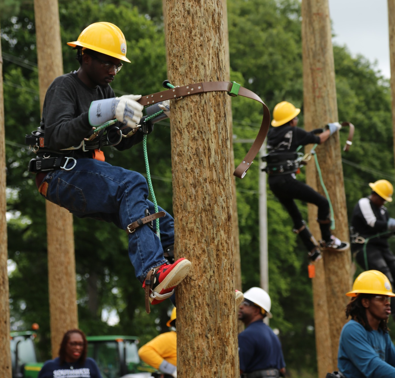 Workers in helmets and safety gear climb tall wooden poles in an outdoor training exercise, showcasing teamwork and focus against a backdrop of trees.