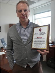 Matthew Lexow standing in his office with a 2025 Jillian Kinzie Award for Recognizing Innovative Leadership for High-Impact Practices plaque in grey cardigan.