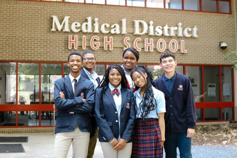 Group of high school students in school uniforms smiling outside a brick building with red doors and windows, standing beneath a sign that reads “Medical District High School,”.
