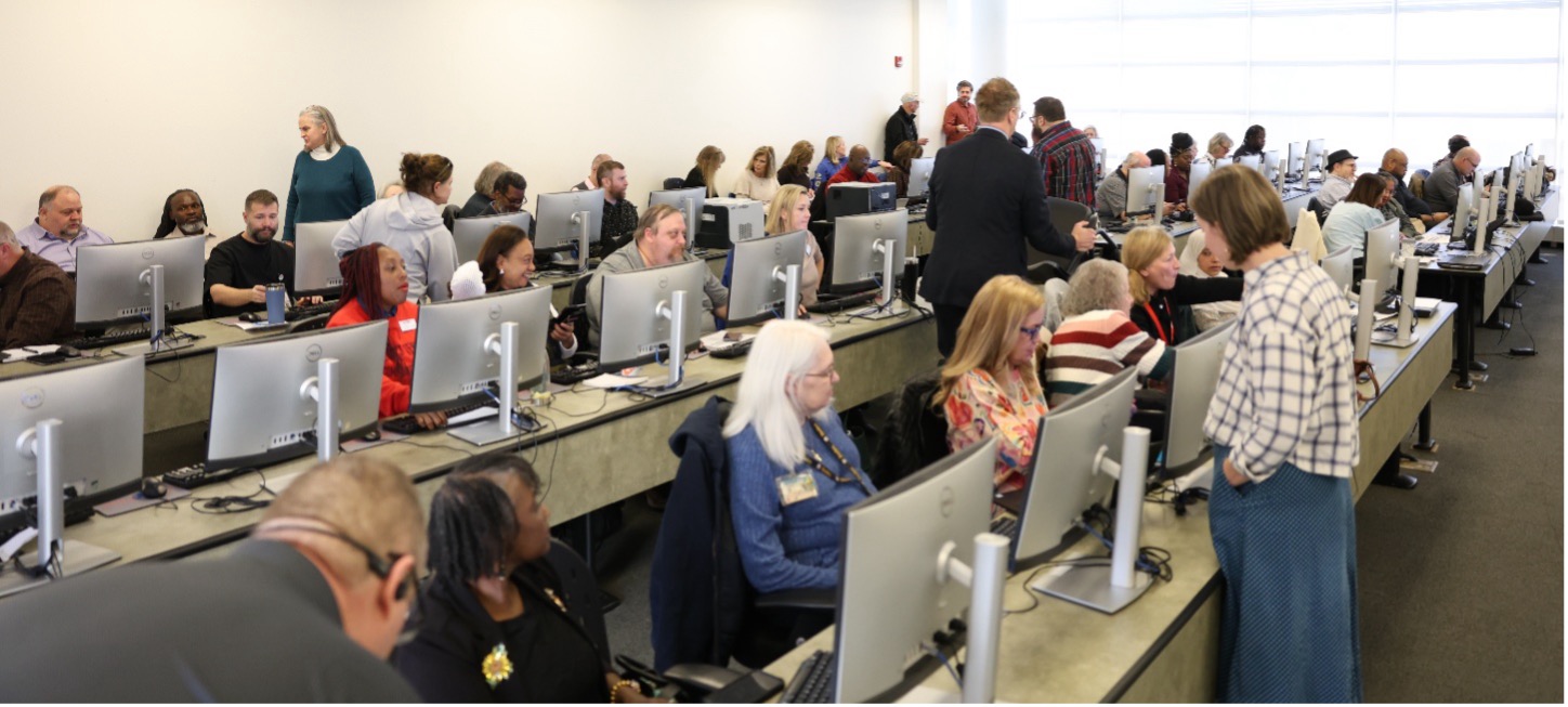 A large group of people in a bright computer lab, each seated at a desktop. Conversations suggest collaboration and a focused, educational atmosphere.